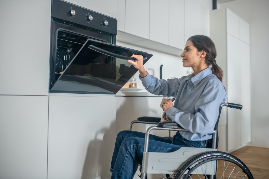 A girl on a wheelchair opening oven in the kitchen while cooking soemthing